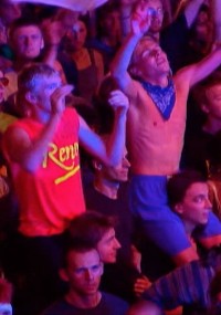 Tłum ludzi na koncercie lub festiwalu, na pierwszym planie widać dwóch młodych mężczyzn; jeden z nich ma na sobie czerwoną koszulkę Reno, drugi jest bez koszulki, ale ma na sobie niebieską bandanę. Obaj wyglądają na podekscytowanych i podnoszą ręce.