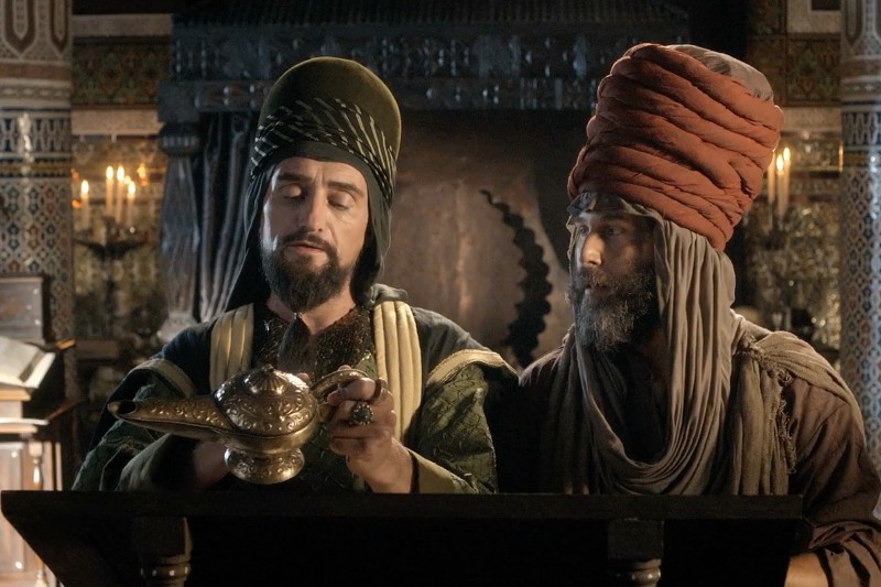 Dwóch mężczyzn w wymyślnych ubraniach w stylu bliskowschodnim stoi w pomieszczeniu. Jeden z nich trzyma dekoracyjną lampę, podobną do lampy Aladyna, podczas gdy drugi patrzy. Ozdobne tło sugeruje scenerię historyczną lub fantastyczną.