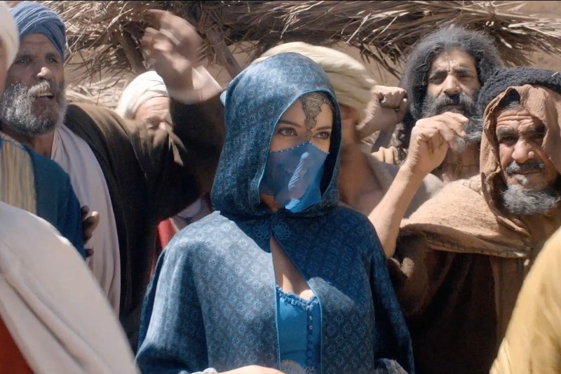 Kobieta w niebieskim wzorzystym kapturze i welonie stoi wśród grupy mężczyzn w tradycyjnych bliskowschodnich strojach, na tle konstrukcji ze słomianymi dachami. Tłum wydaje się ożywiony i ekspresyjny.