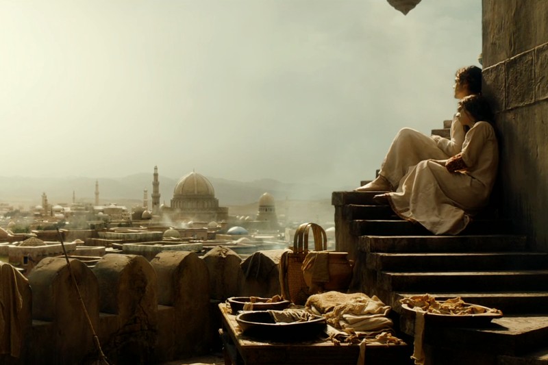 Dwie osoby w jasnych szatach siedzą na kamiennych schodach z widokiem na miasto z kopułami i minaretami. Na pierwszym planie znajduje się stół z jedzeniem i koszami. Scena jest skąpana w ciepłym, miękkim świetle, przywołującym historyczne lub starożytne otoczenie.