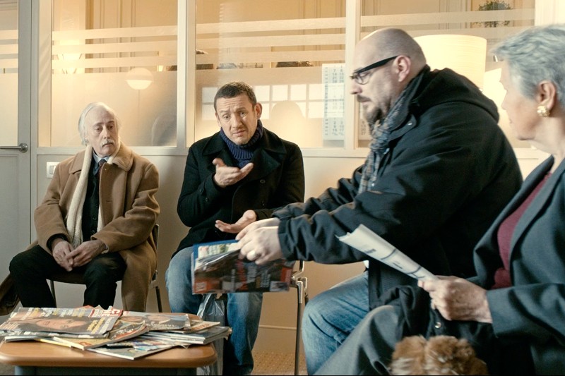 Cztery osoby siedzą w poczekalni z czasopismami na stole. Trzy są zajęte rozmową, podczas gdy jeden, starszy mężczyzna w brązowym płaszczu, patrzy w bok. Miejsce wydaje się być biurem lub kliniką.