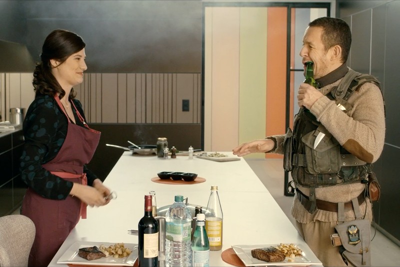 Kobieta w fartuchu uśmiecha się przez kuchenną wyspę do mężczyzny w stroju taktycznym, który pije z butelki. Dwa talerze z jedzeniem i różne butelki ustawione są na białym blacie między nimi.