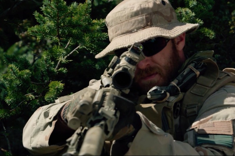 Żołnierz w kamuflażu i czapce moro celuje z karabinu z lunetą, ukrywając się wśród zielonych liści. Na jego mundurze widoczna jest naszywka z amerykańską flagą.