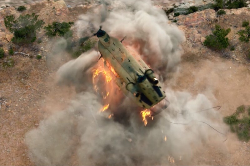 Wojskowy śmigłowiec rozbija się o ziemię, otoczony płomieniami i gęstym dymem, a jego szczątki leżą rozrzucone po skalistym, jałowym terenie.