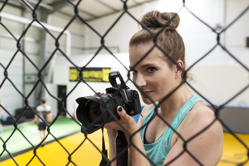 Kobieta w turkusowym podkoszulku trzyma aparat i patrzy przez siatkę ogrodzeniową, przygotowując się do zrobienia zdjęcia w krytym obiekcie sportowym.