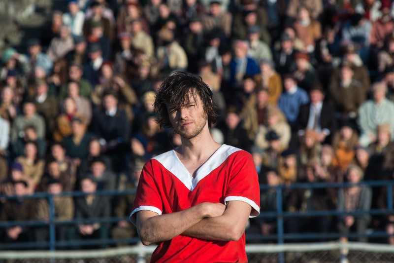 Młody mężczyzna w czerwono-białej koszulce sportowej stoi na boisku ze skrzyżowanymi ramionami i wygląda poważnie, podczas gdy za nim na trybunach stadionu gromadzi się niewyraźny tłum widzów.