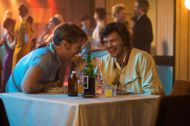 Dwóch mężczyzn siedzi przy stole w ożywionym, ciepło oświetlonym barze lub restauracji, śmiejąc się i pochylając ku sobie. Na stole znajdują się różne butelki i szklanki, a w tle inni ludzie towarzysko rozmawiają.