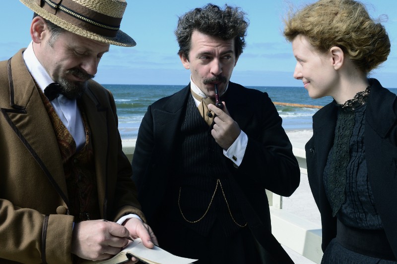 Trzy osoby w ubraniach vintage stoją nad brzegiem morza; jeden mężczyzna pisze na papierze, drugi trzyma fajkę i wygląda na zamyślonego, podczas gdy kobieta się do nich uśmiecha. Niebo jest czyste, a ocean jest w tle.