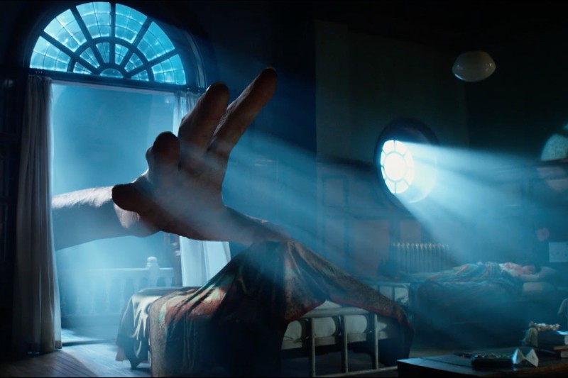 Olbrzymia ręka sięga do nasłonecznionej sypialni z dwoma łóżkami, odsłania koc, a przez duże okrągłe okna wpada błękitne światło, oświetlając kurz i meble w pokoju.