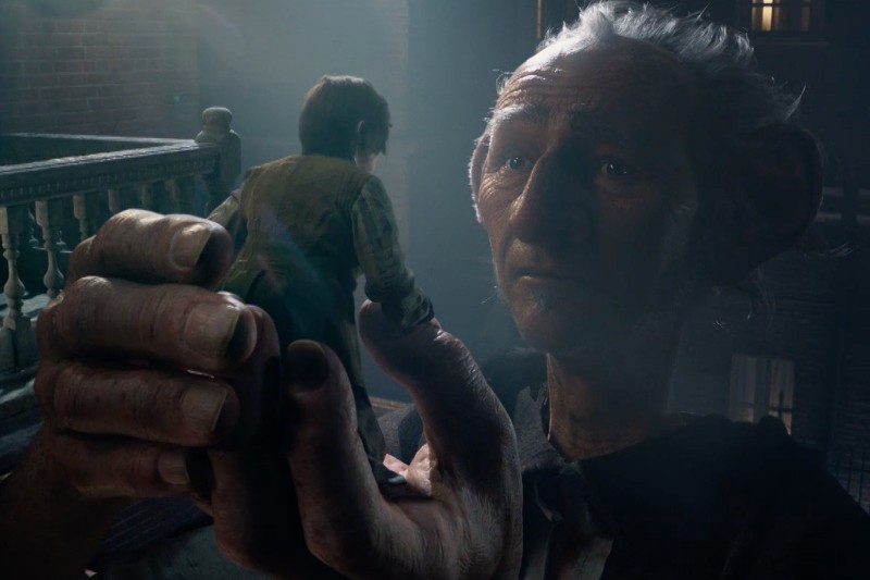 Olbrzymi, łagodnie wyglądający mężczyzna ostrożnie trzyma na ręku małe dziecko na słabo oświetlonym balkonie. W tle widać budynek, a delikatne światło oświetla ich twarze.