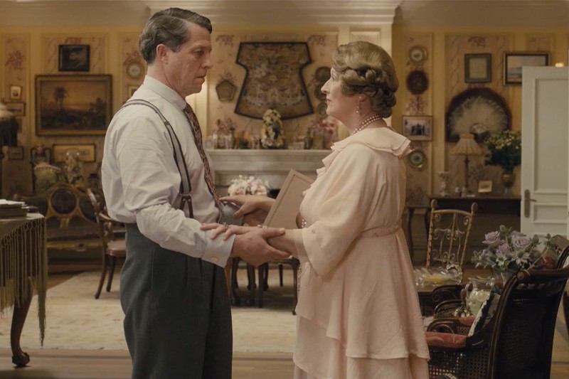 Mężczyzna w białej koszuli i szelkach trzyma za rękę kobietę w jasnoróżowej sukience w bogato zdobionym pokoju pełnym antyków, oprawionych obrazów i dekoracyjnych wachlarzy na ścianach.