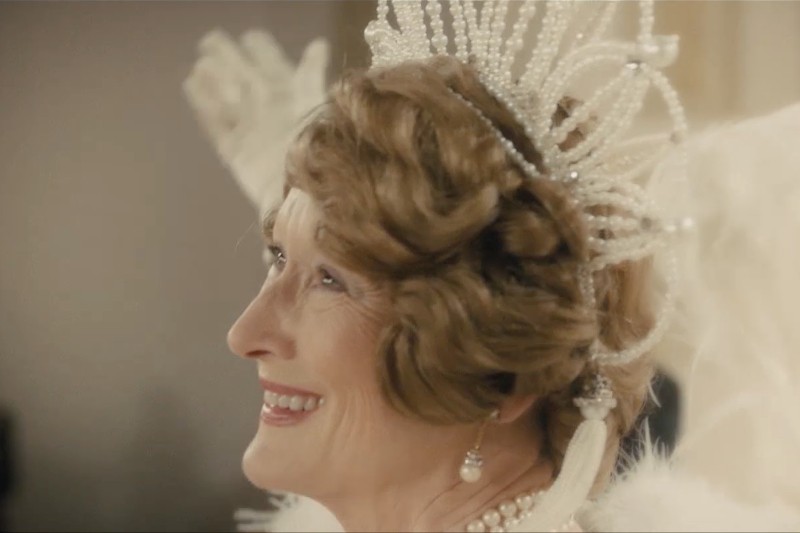 Kobieta o jasnobrązowych włosach uśmiecha się, mając na sobie koralikową, perłową tiarę i pasujące kolczyki, ubrana jest na biało z akcentami z piór i patrzy w bok.