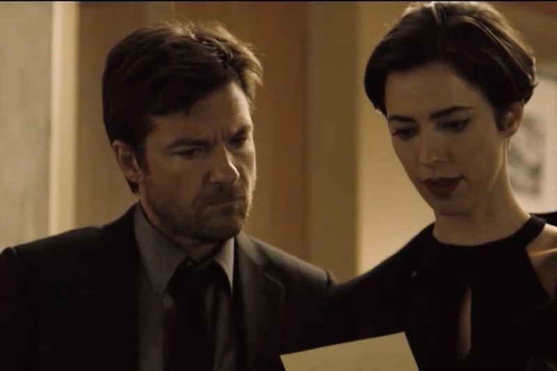 Mężczyzna i kobieta, oboje formalnie ubrani w ciemne ubrania, stoją blisko siebie w pomieszczeniu, uważnie wpatrując się w kartkę papieru, którą trzyma kobieta. Oświetlenie jest słabe i poważne.