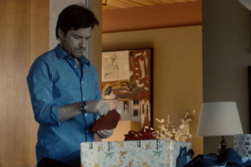 Mężczyzna w niebieskiej koszuli otwiera kartkę z pozdrowieniami obok dużego, zapakowanego prezentu z czerwoną kokardą w ciepło oświetlonym pokoju o nowoczesnym wystroju.