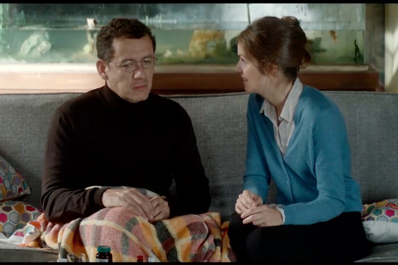 Mężczyzna w okularach i ciemnym swetrze siedzi na kanapie przykrytej kraciastym kocem, a kobieta w niebieskim kardiganie siedzi obok niego, pochyla się i rozmawia z nim.