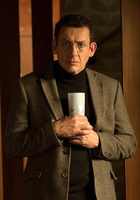 Mężczyzna w okularach i brązowej marynarce na czarnym golfie trzyma w obu rękach biały kubek. Stoi w pomieszczeniu przy łagodnym oświetleniu.