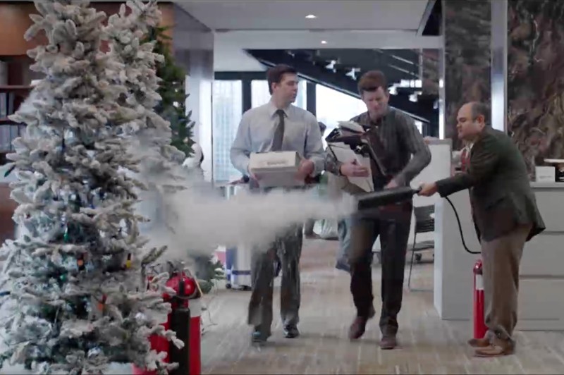 Mężczyzna używa gaśnicy, aby rozpylić sztuczny śnieg na sztucznej choince w biurze, podczas gdy dwóch współpracowników przechodzi obok, niosąc pudełka i urządzenia elektroniczne.