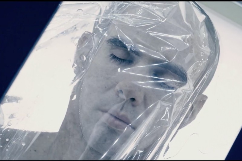 Zbliżenie osoby z zamkniętymi oczami i spokojnym wyrazem twarzy, widzianej przez półprzezroczystą folię plastikową, która częściowo owija się wokół jej głowy. Tło jest jasne i delikatnie oświetlone.