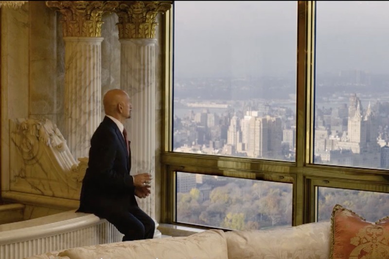 Mężczyzna w garniturze siedzi przy dużym oknie z ozdobnymi kolumnami i w mglisty dzień patrzy na panoramę miasta i park poniżej.