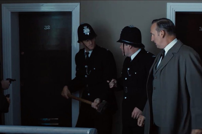 Trzech mężczyzn stoi na korytarzu; dwóch jest ubranych jak brytyjscy policjanci, jeden trzyma młot kowalski, podczas gdy trzeci mężczyzna w garniturze patrzy. Stoją przed drzwiami oznaczonymi numerem 32.