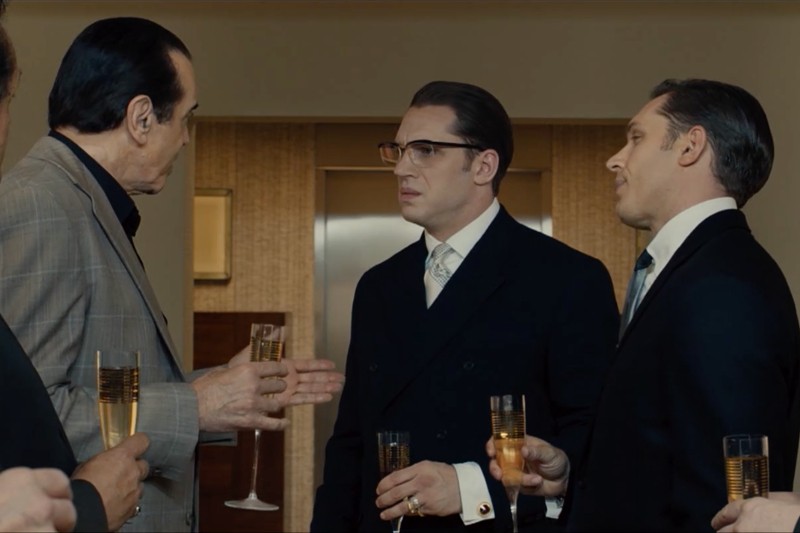 Trzech mężczyzn w garniturach stoi w pomieszczeniu, trzymając kieliszki szampana, zajętych rozmową. Dwóch z nich stoi blisko siebie, podczas gdy trzeci gestykuluje ręką. Sceneria wydaje się być nowoczesnym, dobrze oświetlonym pokojem.