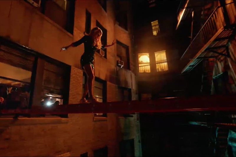 Kobieta w krótkiej sukience pewnie przechadza się nocą wąską belką między budynkami. Okna są ciepło oświetlone, a ludzie obserwują ją z wnętrza.