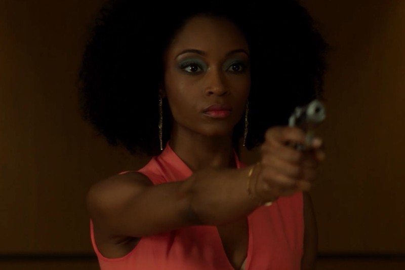 Kobieta o naturalnie kręconych włosach, ubrana w bezrękawnik w kolorze koralowym i długie kolczyki, stoi wewnątrz budynku i celuje pistoletem przed siebie z poważnym wyrazem twarzy.