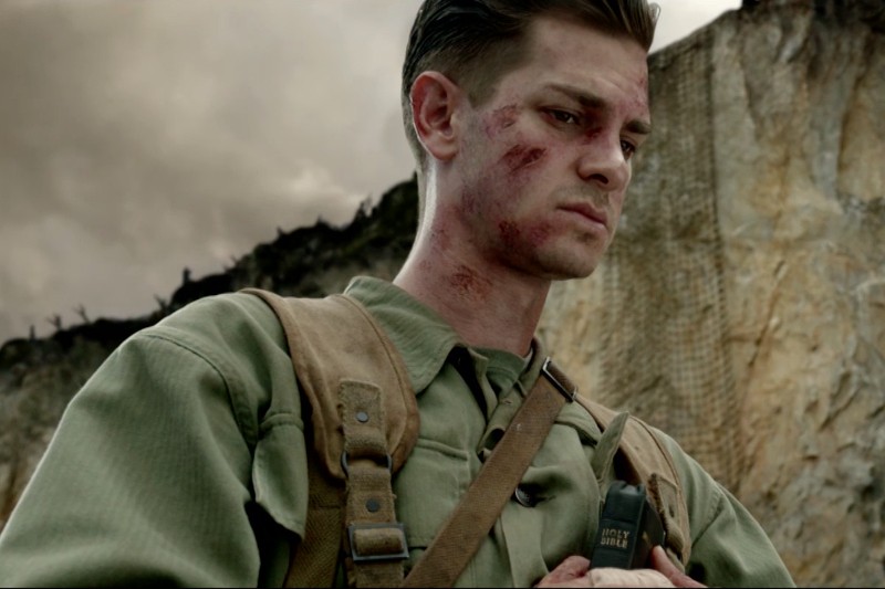 Młody mężczyzna w znoszonym mundurze wojskowym z posiniaczoną twarzą spogląda ponuro w dół, trzymając małą Biblię. Za nim widać postrzępiony, skalisty klif pod zachmurzonym niebem, co sugeruje napięte, rozdarte wojną otoczenie.