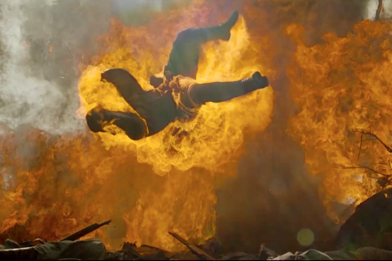 Człowiek zostaje wyrzucony do tyłu w powietrze, otoczony przez duży wybuch pomarańczowych płomieni i dymu po eksplozji, a na ziemi poniżej leżą szczątki.