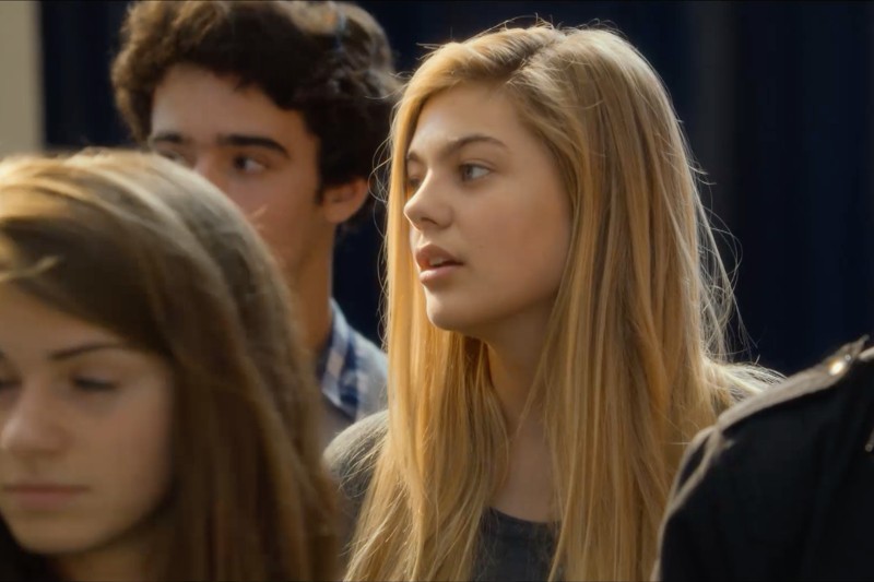 Młoda kobieta z długimi blond włosami stoi w pomieszczeniu wśród grupy ludzi, patrząc w prawo. Dwie inne osoby są widoczne na pierwszym planie, lekko nieostre. Tło jest ciemne i rozmyte.