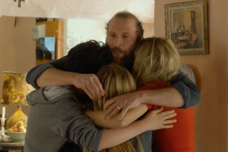 Brodaty mężczyzna obejmuje trzy osoby, dwie z długimi blond włosami i jedną z krótkimi, ciemnymi włosami, w ciepłym, grupowym uścisku w przytulnym, delikatnie oświetlonym pokoju, w tle widoczne są dzieła sztuki i przedmioty codziennego użytku.