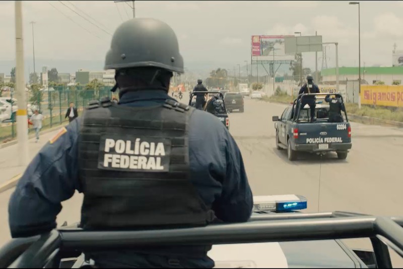 Grupa uzbrojonych funkcjonariuszy meksykańskiej policji federalnej w mundurach jedzie z tyłu pickupów na miejskiej ulicy w ciągu dnia. Główny funkcjonariusz jest widziany od tyłu, ma na sobie hełm i kamizelkę z napisem Policía Federal.