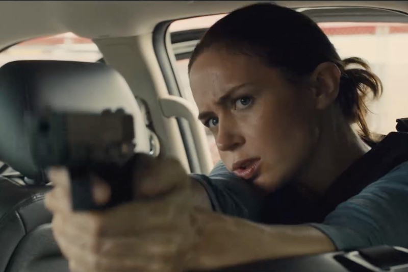 Kobieta z brązowymi włosami związanymi do tyłu intensywnie mierzy z broni z wnętrza samochodu, wydaje się skupiona i czujna.