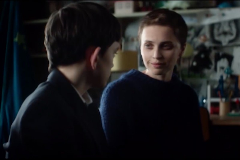 Kobieta i chłopiec siedzą w pomieszczeniu, zwróceni do siebie twarzą w twarz i delikatnie się uśmiechają. W tle widać półki, papiery i różne przedmioty, co sugeruje osobiste lub domowe otoczenie.