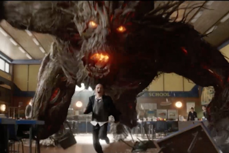 Osoba w formalnym stroju ucieka w strachu, gdy wielki, ognistooki, przypominający drzewo potwór z otwartą paszczą goni ją w kafeterii lub szkolnej stołówce. Stoły i krzesła są przewrócone w chaosie.