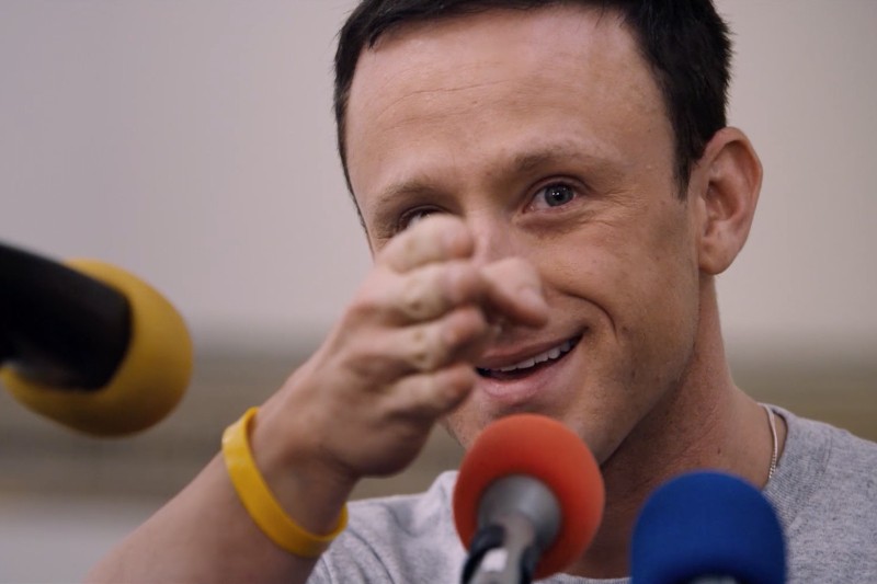 Mężczyzna z krótkimi brązowymi włosami, ubrany w szarą koszulę i żółtą opaskę, uśmiecha się, gestykulując lewą ręką. Przed nim znajdują się trzy mikrofony z kolorowymi osłonami.