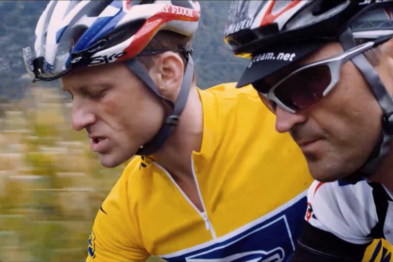 Dwóch rowerzystów w kaskach i okularach przeciwsłonecznych jedzie blisko siebie. Rowerzysta z przodu ma żółtą koszulkę, a ten z tyłu białą, czarną i czerwoną. Tło jest rozmyte, zielone.