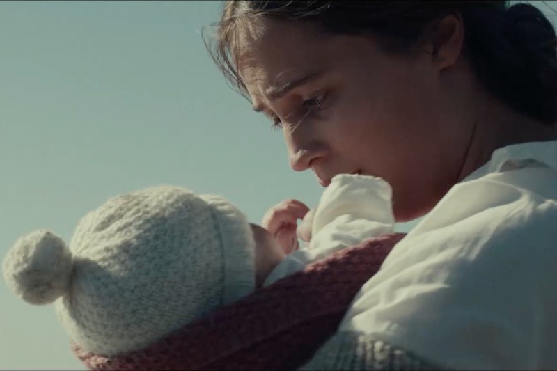 Kobieta w białej bluzce trzyma na rękach dziecko owinięte w wełnianą czapkę i kocyk, patrząc na nie zatroskanym wzrokiem pod czystym niebem.