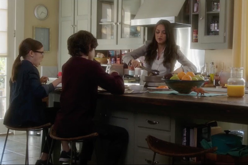 Kobieta serwuje jedzenie dwójce dzieci siedzących przy kuchennej wyspie. Kuchnia jest jasna, na blacie leżą owoce, sok i różne rzeczy. Kobieta wydaje się przygotowywać śniadanie.