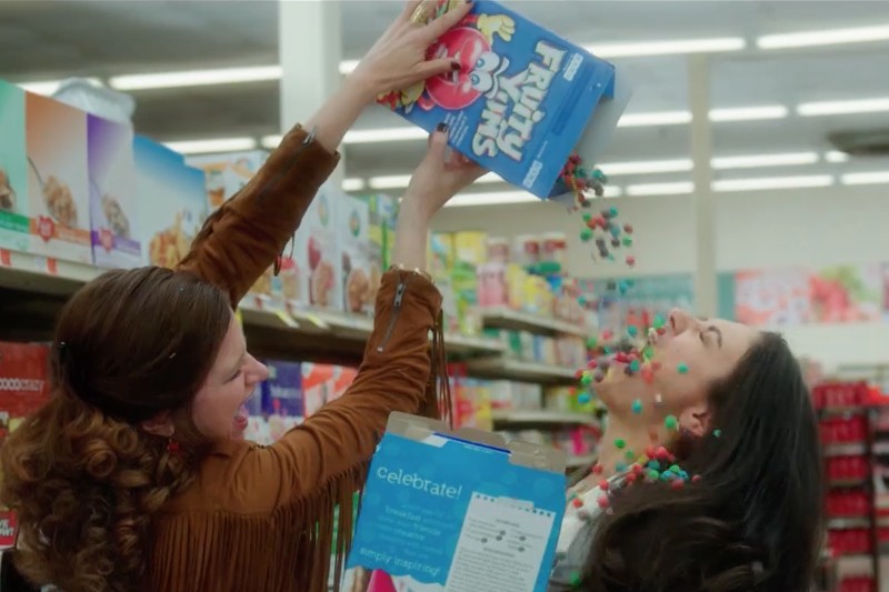 Dwie kobiety w sklepie spożywczym śmieją się i żartobliwie wysypują sobie na siebie zawartość pudełka kolorowych płatków śniadaniowych Froot Loops, przy czym kawałki płatków wysypują się i latają w powietrzu.