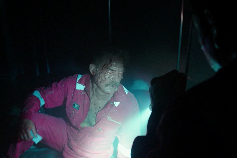 Mężczyzna w czerwonym kombinezonie z zakrwawioną twarzą siedzi na ziemi w słabym oświetleniu, oświetlany latarką trzymaną przez niewidoczną osobę na pierwszym planie.