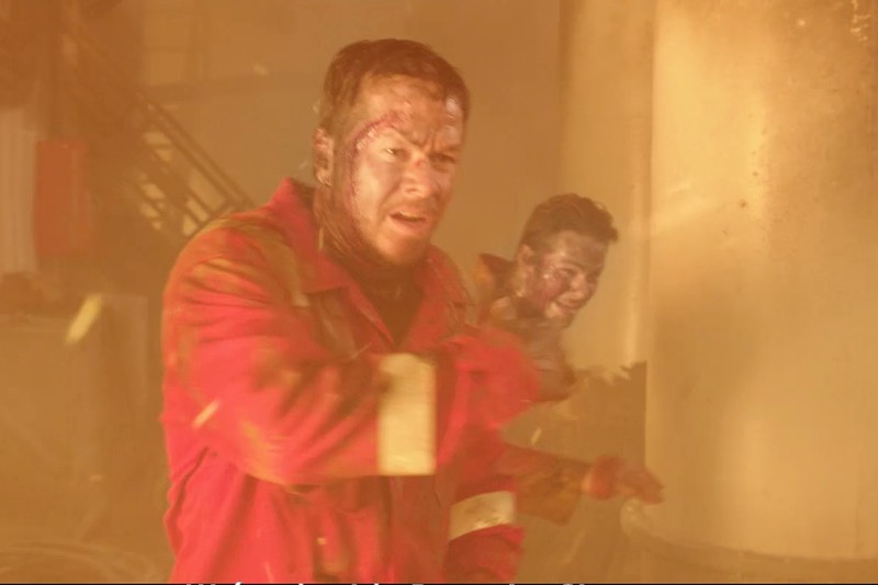 Dwie osoby w czerwonych kombinezonach ochronnych, pokryte brudem i popiołem, poruszają się w zadymionym, chaotycznym środowisku przemysłowym z pomarańczowo-żółtym oświetleniem, co sugeruje sytuację awaryjną lub katastrofę.