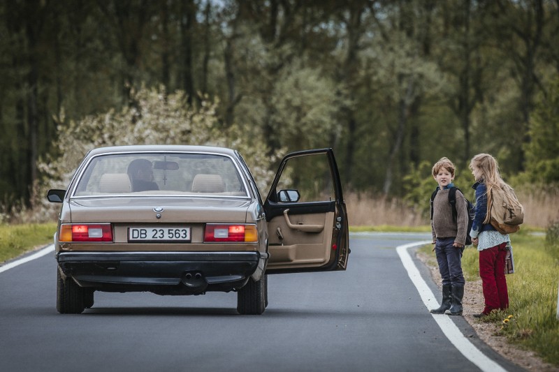 Zaparkowane srebrne BMW z otwartymi drzwiami kierowcy stoi na wiejskiej drodze. Dwoje dzieci, chłopiec i dziewczynka, stoją na poboczu drogi obok samochodu, otoczeni drzewami i trawą.