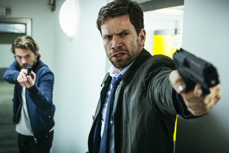 Dwóch mężczyzn w kurtkach stoi na korytarzu, celując pistoletami do przodu z poważnymi minami, jakby stawiali czoła zagrożeniu. Korytarz jest jasno oświetlony, a na ich twarzach widać napięcie.