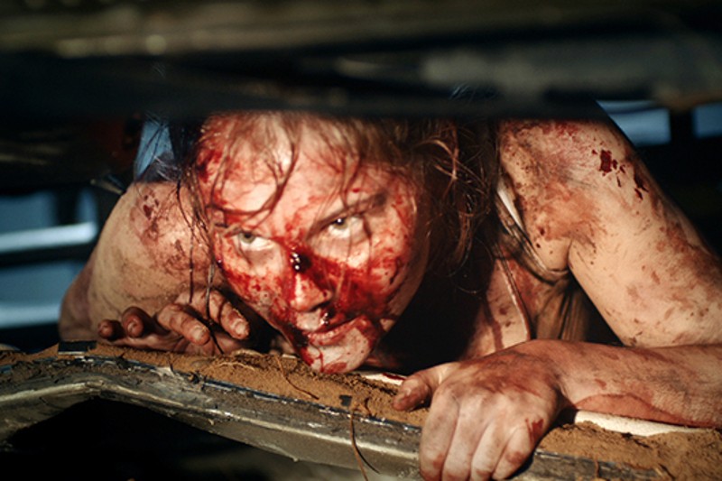 Człowiek z zakrwawioną twarzą i ciałem czołga się po ziemi, patrzy w górę z wyrazem zdecydowania, otoczony gruzami i słabym oświetleniem.