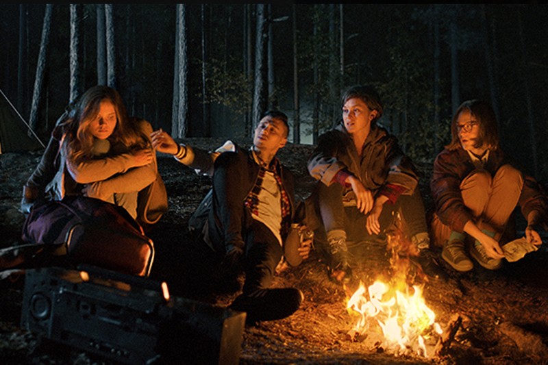 Czwórka młodych ludzi siedzi nocą przy ognisku w ciemnym lesie, otulona kurtkami. Wygląda na to, że rozmawiają i cieszą się swoim towarzystwem, a na ziemi w pobliżu leży radio. W tle widać drzewa.
