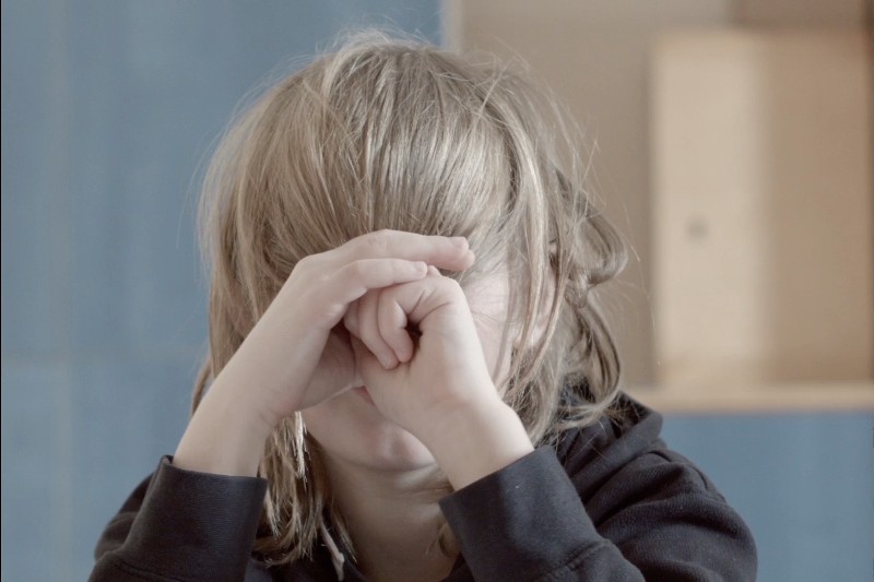 Dziecko z długimi, jasnobrązowymi włosami zakrywającymi twarz, składa ręce przed nosem i ustami, opiera łokcie na stole, ubrane jest w ciemną bluzę, znajduje się w łagodnie oświetlonym pokoju.