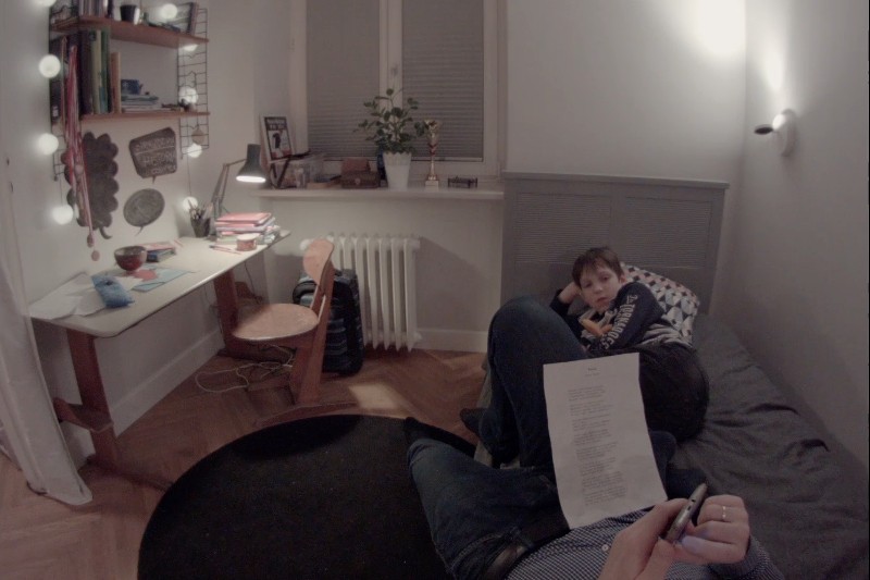 Przytulna sypialnia z biurkiem i półkami po lewej stronie, osoba leżąca na łóżku trzymająca papiery na pierwszym planie oraz małe dziecko siedzące na łóżku i patrzące w stronę aparatu.