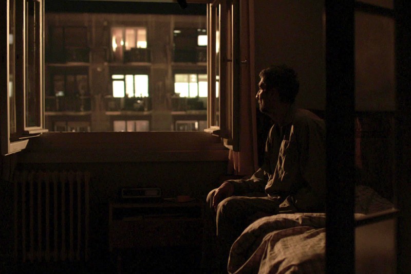 Mężczyzna siedzi na skraju łóżka w słabo oświetlonym pokoju i patrzy przez otwarte okno na budynek mieszkalny z oświetlonymi nocą oknami.