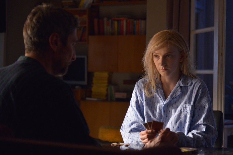 Kobieta w piżamie siedzi przy stole, trzymając karty do gry, i poważnie patrzy na mężczyznę siedzącego naprzeciwko niej w słabo oświetlonym pomieszczeniu z regałami z książkami w tle.